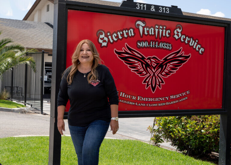 Terry Quinones, owner of Sierra Traffic Service, wears a black shirt and jeans standing in front of a red, white, and black sign reading "Sierra Traffic Service" and their contact information.