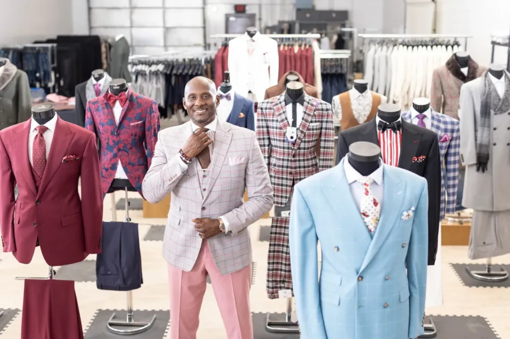 Montee adjusts his tie surrounded by suits displayed in his workspace.