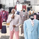 Montee adjusts his tie surrounded by suits displayed in his workspace.