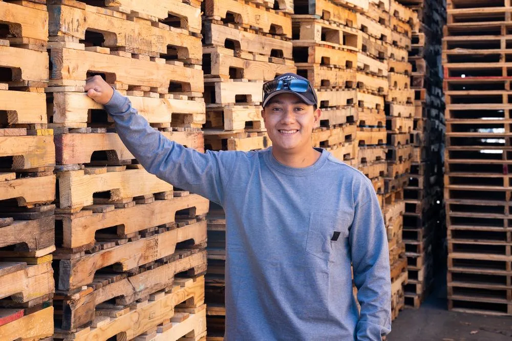 Juan Jr. in front of tall stacks of pallets at Johnny’s Pallet Woodworks which was aided by small business financing from a SBA 504 loan with CDC Small Business Finance.