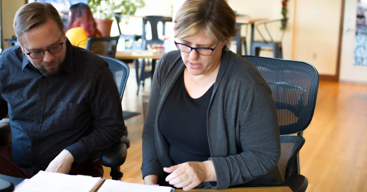 A woman in sits at a desk with a colleague, reviewing financial projections.